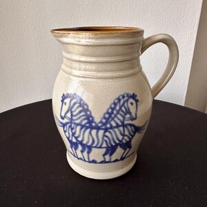 May include: A beige ceramic pitcher with a handle and a brown rim. The pitcher features a blue illustration of two zebras facing each other. The pitcher is a decorative item for the kitchen or dining room.