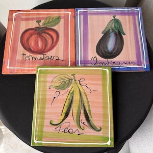 May include: Three square wooden plaques with painted images of vegetables. One shows a red tomato with the word "tomatoes". Another depicts a purple eggplant with the word "Ombergines". The third shows green peas with the word "Peas".