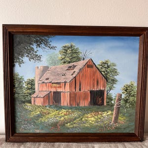 May include: Oil painting of a weathered red barn with a silo, set amidst lush green trees and a field of wildflowers. The artwork is framed in a dark brown wooden frame, capturing a rural landscape scene.