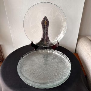 May include: Clear glass plates with a textured, leaf-like pattern. One plate is displayed on a dark wooden stand, while a stack of similar plates rests below. The plates have a scalloped edge.