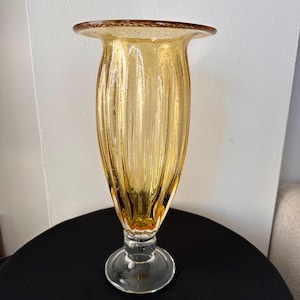 May include: A tall, amber-colored glass vase with a flared top and a clear, circular base. The vase has vertical ribbed detailing and a slightly textured surface. The rim of the vase has a darker amber hue.