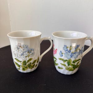 May include: Two white ceramic mugs with floral designs. One mug features blue flowers, the other has pink and blue flowers. Both mugs have green leaves and textured handles.