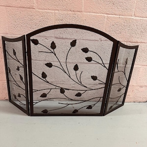 May include: A dark brown, three-panel fireplace screen. The screen features a mesh design with decorative metal leaf and vine accents. The screen is designed to stand on its own and protect the area around a fireplace.