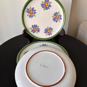 May include: Set of three ceramic plates. Each plate features a white center with a floral design in blue, red, and yellow. The rims are green. The underside of one plate has the inscription "C. Peru".