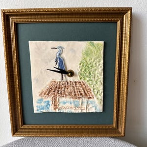 May include: A square wall clock featuring a textured ceramic tile with a blue heron standing on a thatched roof, set against a dark green mat and a gold-toned ornate frame. The clock has two black hands centered on the heron's body.