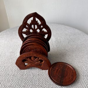May include: A set of intricately carved wooden coasters and a matching holder. The coasters are round, with detailed floral patterns and a rich brown finish. The holder features a decorative openwork design. Each coaster is approximately 4 inches in diameter.