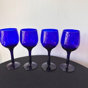 May include: Four cobalt blue wine glasses with a rounded bowl and a long stem. The glasses are arranged in a row on a dark surface, with a white wall in the background. The glasses are made of glass.