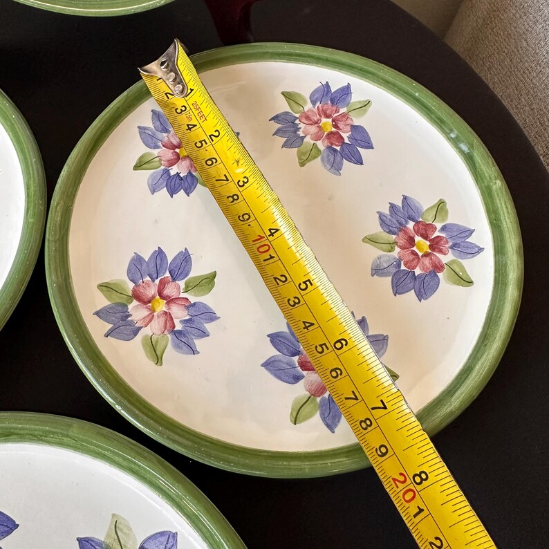 May include: White ceramic plate with a green rim and floral design. The plate features blue and pink flowers. A measuring tape shows the plate is approximately 8 inches in diameter.