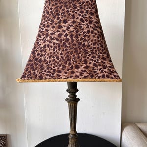 May include: A table lamp featuring a pyramid-shaped lampshade with a brown and black leopard print pattern and gold trim. The lamp has a dark, weathered metal base with a fluted column design and a small, decorative finial at the very top.