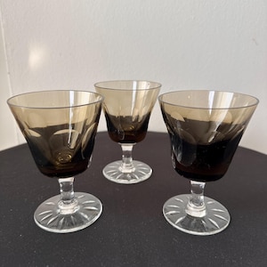 May include: Three vintage brown-tinted stemmed glasses. The glasses have a flared bowl design and sit on a clear, etched base. The stems are clear, and the glasses are arranged on a dark surface. The glasses are likely for serving wine or other beverages.