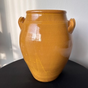May include: A glazed, mustard-yellow ceramic jar with two curved handles. The jar has a rounded body and a slightly flared rim. The surface is smooth and reflective, with subtle variations in color. A decorative storage container.