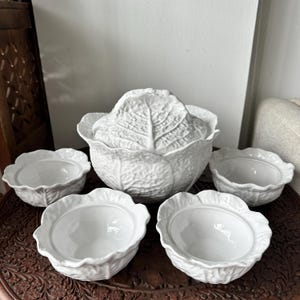 May include: A white ceramic serving set featuring a large lidded tureen and four matching bowls. Each piece is molded with a detailed cabbage leaf texture, including prominent veins and ruffled edges, creating a cohesive botanical design.