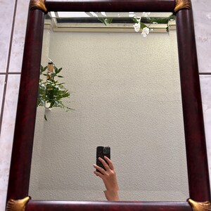 May include: A rectangular wall mirror featuring a dark reddish-brown faux wood grain frame. The four corners of the frame are accented with decorative gold-colored metal caps that display a raised, fan-like shell pattern on each corner.