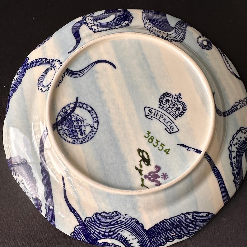 Puede incluir: Plato de cer&aacute;mica blanco y azul con dise&ntilde;o n&aacute;utico. El plato presenta tent&aacute;culos de pulpo azules alrededor del borde y un dise&ntilde;o central con un barco y el texto "ASSOCIATION". El plato tambi&eacute;n tiene el texto "S.H.& Co." y el n&uacute;mero "38354".