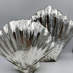 May include: Two decorative shell-shaped objects with a reflective, silver-toned mercury glass finish. Each piece features a ribbed, fan-like texture with a distressed, speckled appearance across the surface, creating a vintage metallic aesthetic.