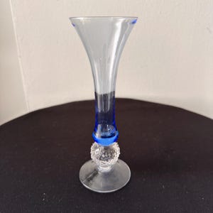 May include: A clear glass vase with a flared top and a blue base. The vase has a decorative, textured ball near the base and a round, clear base. The vase is approximately 8 inches tall.