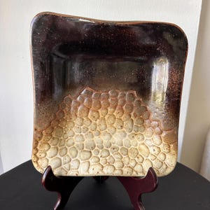May include: Square ceramic dish with a textured, honeycomb-like pattern in shades of brown and cream. The dish has a dark brown border and is displayed on a wooden stand. The dish is likely handmade.