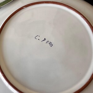 May include: A white ceramic plate with a brown rim. The plate has the handwritten text "C. PERS" in black ink. The plate is likely handmade and suitable for serving food.
