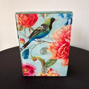 May include: A decorative three-drawer chest with a floral and bird design. The chest features a light blue background with red roses, orange peonies, and a colorful bird. Each drawer has a clear, crystal-like knob.