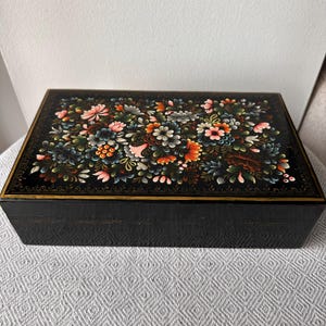 May include: A rectangular black wooden box with a floral design on the lid. The flowers are in shades of pink, orange, blue, and green. The box has a gold trim and a textured exterior.