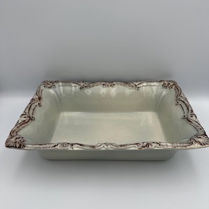 May include: A rectangular ceramic baking dish with a glossy off-white interior and a scalloped rim. The edges feature an ornate, raised relief pattern highlighted with a dark brown, weathered finish that contrasts against the light base.