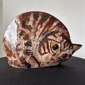 May include: A ceramic cat figurine in a resting pose. The cat has brown and tan striped fur, with large yellow eyes and a pink nose. The figurine is on a black surface.