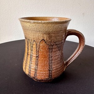 May include: A handmade ceramic mug with a textured surface. The mug features a brown and tan color scheme with vertical black lines and a curved handle. The mug is approximately 4 inches tall and 3 inches wide.