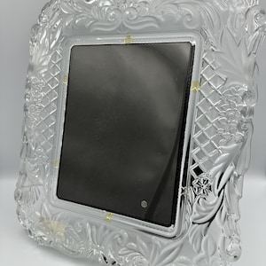 May include: A square picture frame made of clear glass with a decorative border featuring embossed floral patterns and a lattice design. The frame has a central black rectangular insert and a polished, reflective surface with intricate relief details.