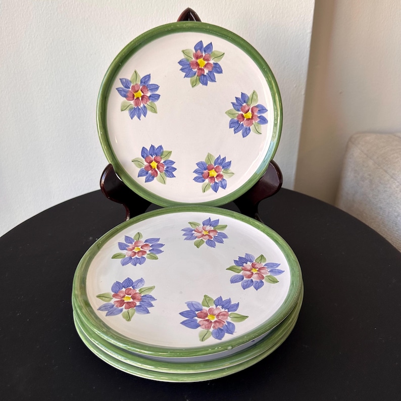May include: Set of white ceramic plates with a green rim and hand-painted floral design. Each plate features six flowers with blue, pink, and yellow petals. The plates are stacked and displayed on a dark surface.