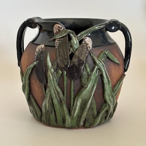 May include: A ceramic vase with a dark brown exterior and black rim, featuring sculpted cattails and green reeds. The vase has two black handles and a textured surface, creating a rustic, nature-inspired design.