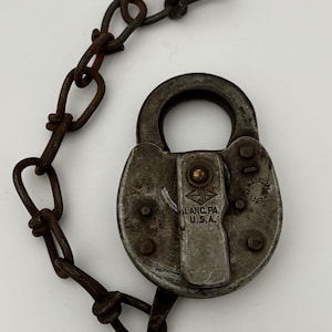 Puede incluir: Un candado antiguo y desgastado con una cadena. El candado es de color plateado opaco y tiene el texto "LANC. PA. U.S.A." y "MADE IN U.S.A.". La cadena y el candado muestran signos de edad y desgaste.