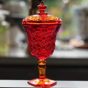 Puede incluir: Un cuenco de cristal rojo vibrante con tapa, que presenta un patrón detallado de estrellas y diamantes prensados en el cuerpo y la cubierta. La pieza se asienta sobre una base de pedestal con pie hexagonal, mostrando un tono carmesí intenso.