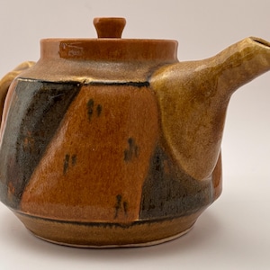 Puede incluir: Una tetera de cerámica con tapa y asa. La tetera presenta un diseño geométrico con colores marrón, negro y beige. El pico es curvo y el asa es redondeada. La tetera se utiliza probablemente para preparar té.