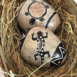 May include: Two decorative stone eggs with carved designs of a turtle and a lizard, resting in a woven basket filled with dried grass. The eggs are beige with black and brown accents.