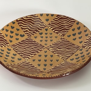 Puede incluir: Un plato decorativo redondo con borde marrón y centro beige. El plato está dividido en cuadrados y triángulos con patrones alternos de líneas onduladas marrones, pequeños corazones negros y cuadrados beige. El plato tiene una apariencia rústica y hecha a mano.