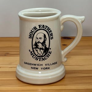 May include: A cream-colored ceramic mustache mug featuring a black graphic of a man with a mustache inside an oval frame. The text reads Your Father's Mustache, Greenwich Village, New York. The mug has a handle and a built-in mustache guard shelf.