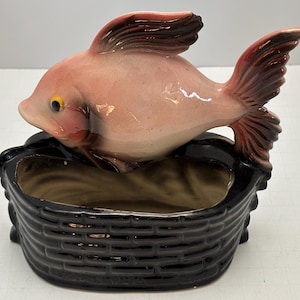 May include: A ceramic fish ornament in a black basket-style planter. The fish is pink with dark red fins and tail, and a yellow eye. The planter has a basket weave design.