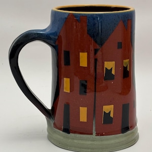 Puede incluir: Una taza de cerámica con asa, con un diseño de edificios rojos con puertas y ventanas negras, y luces amarillas en las ventanas. La parte superior de la taza es azul y la base es verde.