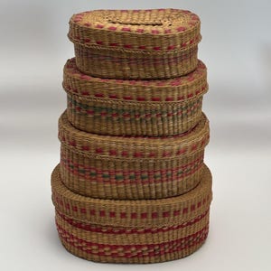 May include: A stack of five oval-shaped woven baskets with lids. The baskets are made of natural-colored straw and feature red and green decorative bands. The lids fit snugly on top, creating a cohesive set for storage or display.