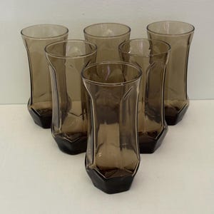 May include: Set of six vintage brown tinted drinking glasses. Each glass has a flared, faceted design and a dark brown, geometric base. The glasses are arranged in a triangular formation, showcasing their unique shape and color.