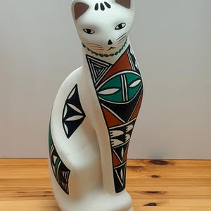 May include: A white ceramic cat figurine featuring a matte finish and painted geometric patterns in black, terracotta, and green. The cat has a stylized face with black markings, a green beaded collar design, and a tall, slender, seated posture.