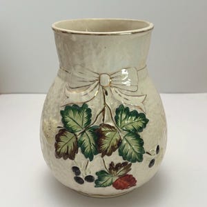 May include: A cream-coloured ceramic vase with a textured surface, featuring a raised relief design of a bow tied around the neck. The body is decorated with painted green and brown leaves, small dark berries, and a single red berry, with gold trim.
