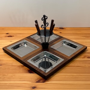 May include: A square wooden serving tray featuring four recessed stainless steel square bowls. In the center stands a black metal rack with a decorative scrollwork top, designed to hold four small utensils with dark brown wooden handles.