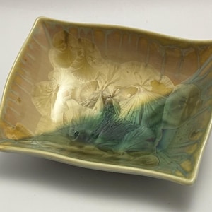 May include: A rectangular ceramic dish with a unique glaze. The dish features a blend of colors, including beige, green, and yellow, creating a marbled effect. The edges are slightly curved, and the overall design is artistic and decorative.