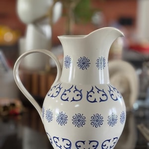 May include: A white ceramic pitcher with a curved handle and spout. The pitcher is decorated with a repeating pattern of blue snowflake and scroll designs. The pitcher is a decorative item.