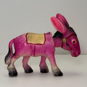 May include: A vintage pink donkey figurine with a saddle. The donkey has black and gray hooves and a black mane. The ears and face are a darker shade of pink. The saddle is tan with a gold trim. The donkey is a decorative item.