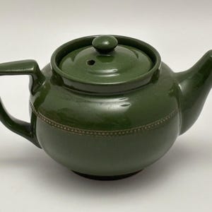 May include: A green ceramic teapot with a matching lid and handle. The teapot has a rounded body with a decorative band around the middle. The spout and handle are curved. The lid has a small knob on top.