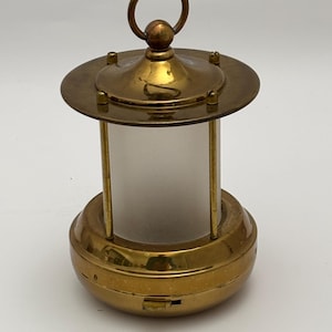 May include: A small, vintage brass lantern with a frosted glass cylinder. The lantern has a circular base and top, connected by vertical brass bars. A small loop is at the top for hanging. The lantern is likely for decorative purposes.