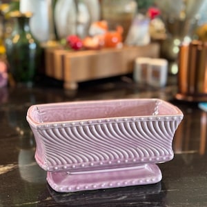 May include: A rectangular, light pink ceramic planter featuring a glossy finish, a raised beaded border along the top and base edges, and a diagonal, wavy, embossed texture across the sides. The planter sits on a small, integrated pedestal base.