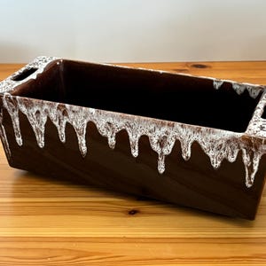 Puede incluir: Una jardinera rectangular de cerámica marrón con un diseño de esmalte blanco goteado. La jardinera tiene dos asas rectangulares recortadas. El interior de la jardinera es marrón oscuro. Está sobre una superficie de madera.